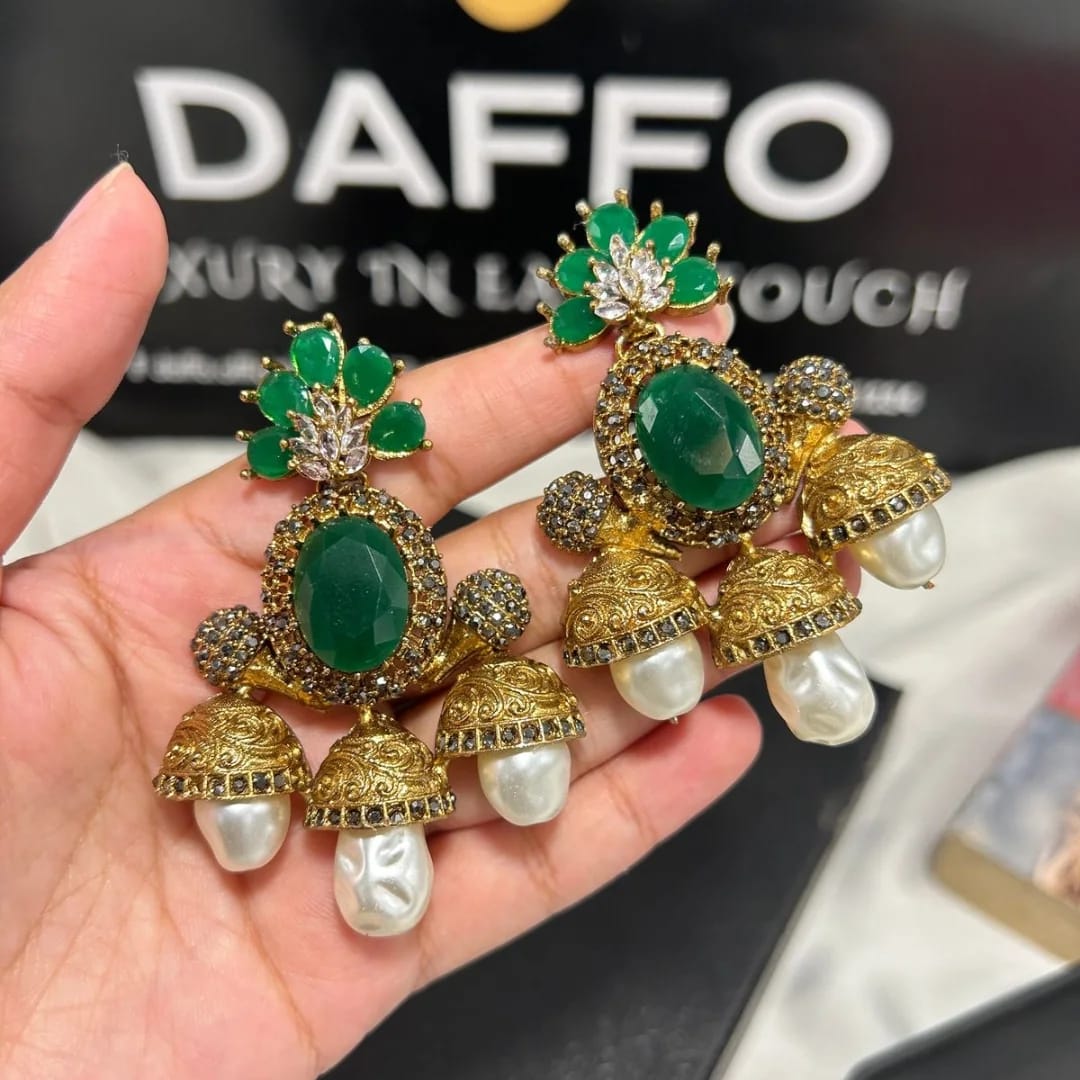 Ornate earrings with green stones and pearls held in a hand, with 'DAFFO' branding in the background.